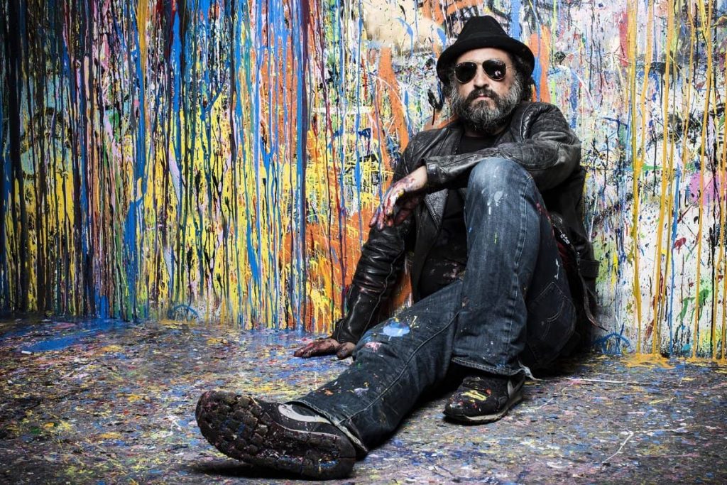 a man sitting in front of a wall which is painted all over with spray paint. He is wearing a black hat and a beard. The man is Thierry Guetta, the artist Mr. Brainwash.